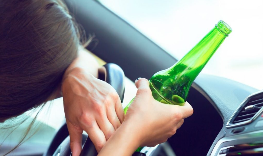 Woman resting head on steering wheel while holding partially empty beer bottle; image by fabrikasimf, via Freepik.com.